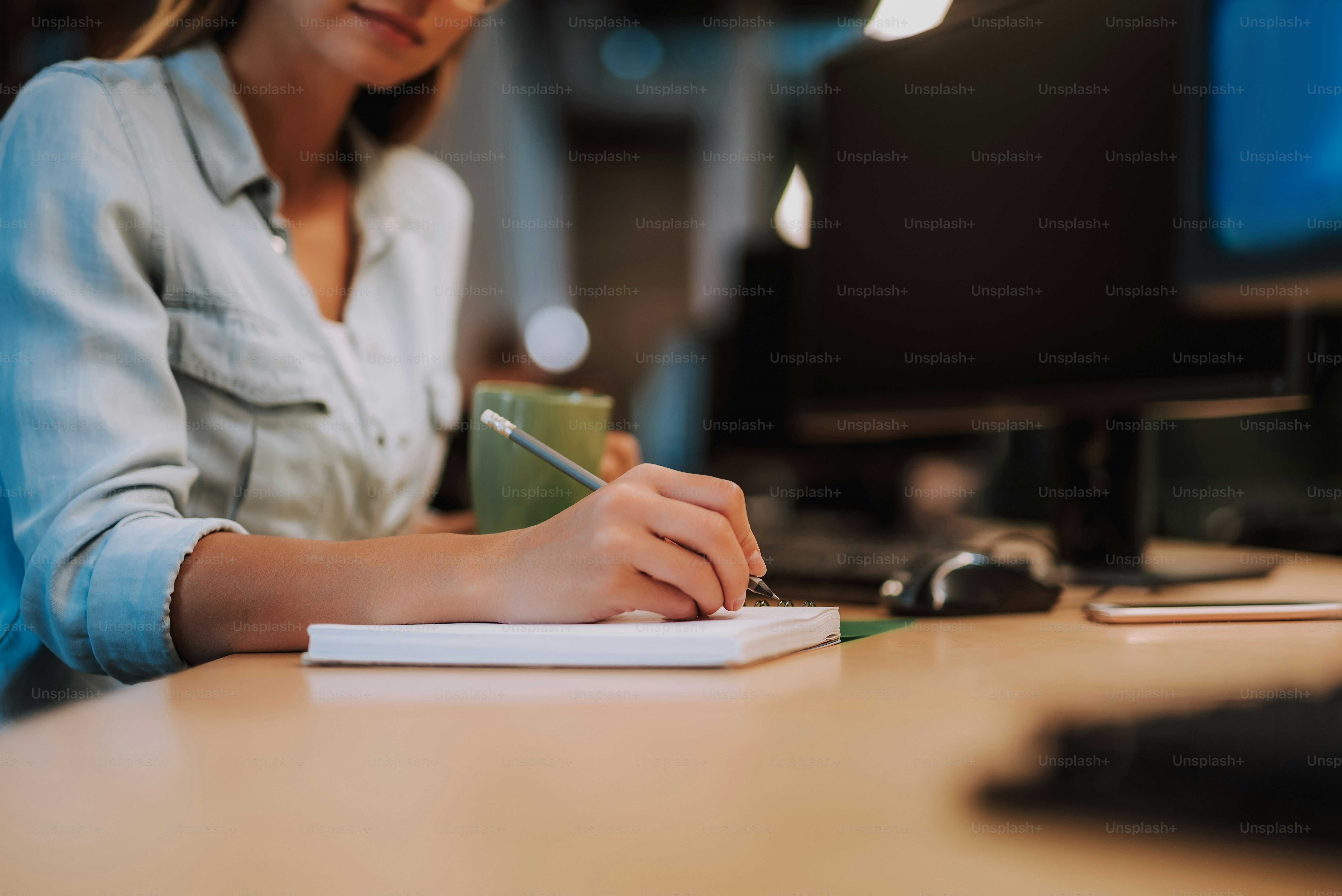 Cropped portrait of lovely girl making notes and holding cup of coffee while sitting at office desk. Focus on woman hand