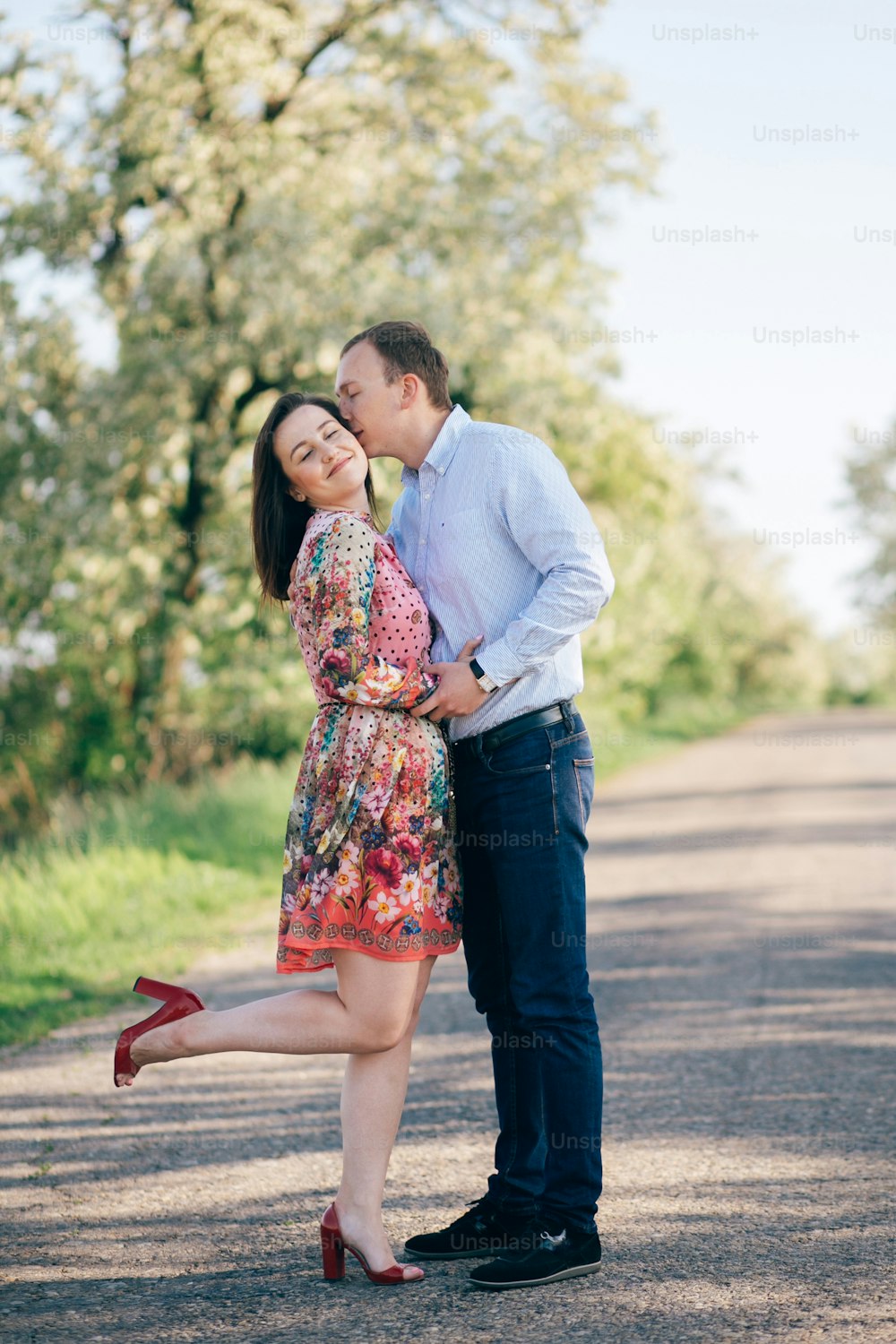 Beautiful young couple sensual hugging and kissing on road in sunshine among spring field and trees. Happy family in love embracing in sunlight. Romantic joyful moments. Space for text