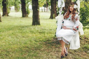 公園でおとぎ話のスイングに刺繍された白いドレスを着た美しい妊娠中のブルネットの女性。母性の優しい瞬間。テキスト用のスペース
