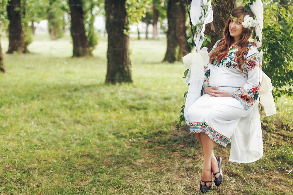 Beautiful pregnant brunette woman in embroidered white dress on fairytale swing in park. tender moment of motherhood. space for text