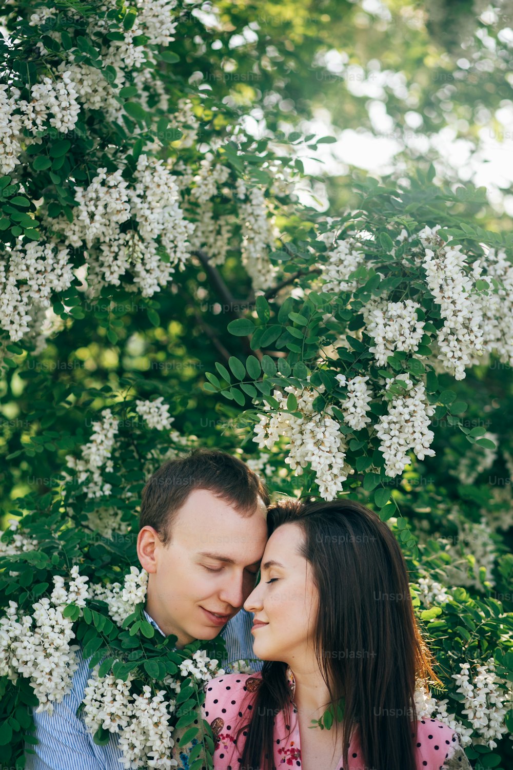 Hermosa pareja joven abrazada suavemente en hojas verdes y flores blancas en el jardín de primavera bajo el sol. Familia feliz abrazada en acacia floreciente a la luz del sol. Momentos románticos.