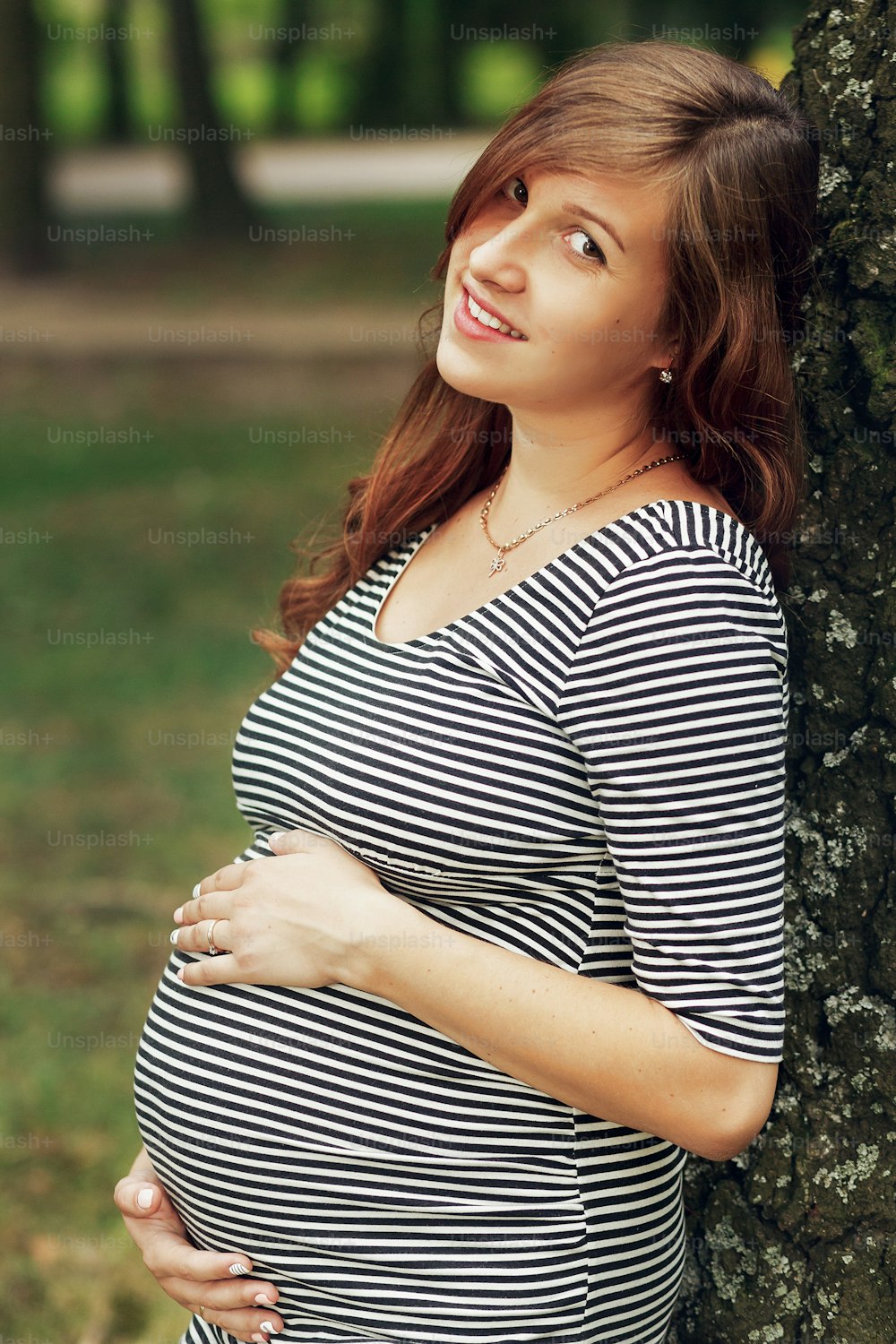 Beautiful brunette pregnant woman in casual clothes, holding belly while posing in park. gentle sweet moment of future motherhood