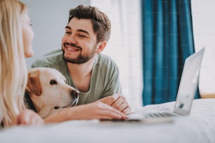 쾌활한 수염을 기른 남자가 침대에서 강아지와 함께 쉬고 있는 동안 여자 친구를 보고 있다