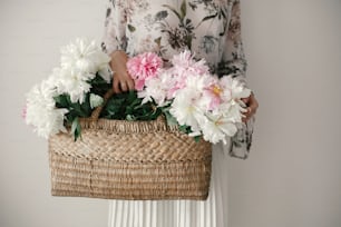 Boho girl holding pink and white peonies in rustic basket. Stylish hipster woman in bohemian floral dress gathering peony flowers. International Womens Day.  Wedding decor and arrangement
