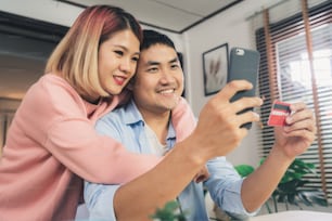 Young Asian family couple using smartphone discussing news or doing online shopping sitting together on desk at home weekend together, serious man and woman browsing web searching for new sale offers.