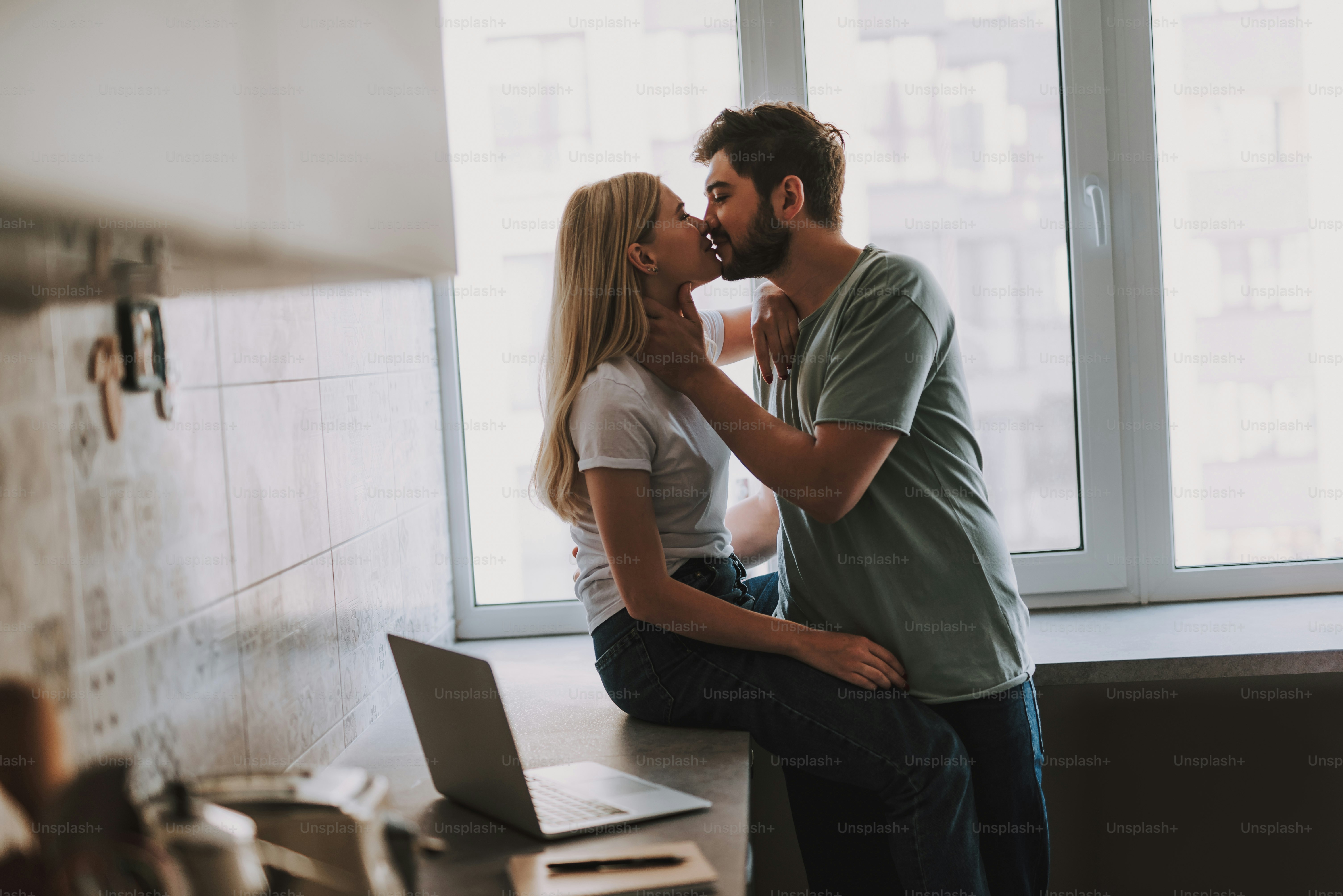 Side view of attractive girl with long blond hair sitting on tabletop near the open laptop and bearded man standing nearby and kissing her