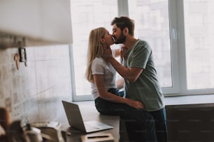 긴 금발 머리를 가진 매력적인 소녀가 열린 노트북 근처의 탁상에 앉아 있고 수염을 기른 남자가 근처에 서서 그녀에게 키스하는 측면 모습