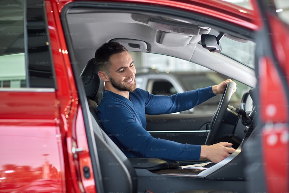 Handsome brunette man sitting in red car with opened door. Attractive guy in blue sweater on driver's seat in new auto holding one his hand on stearing wheel. Young driver looking for something.