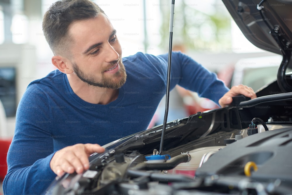 Close up of attractive happy man leaning on opened hood. Young customer in blue sweater choosing car in auto salon. Brunette driver with beard checking engine of new vehicle and squatting near it.