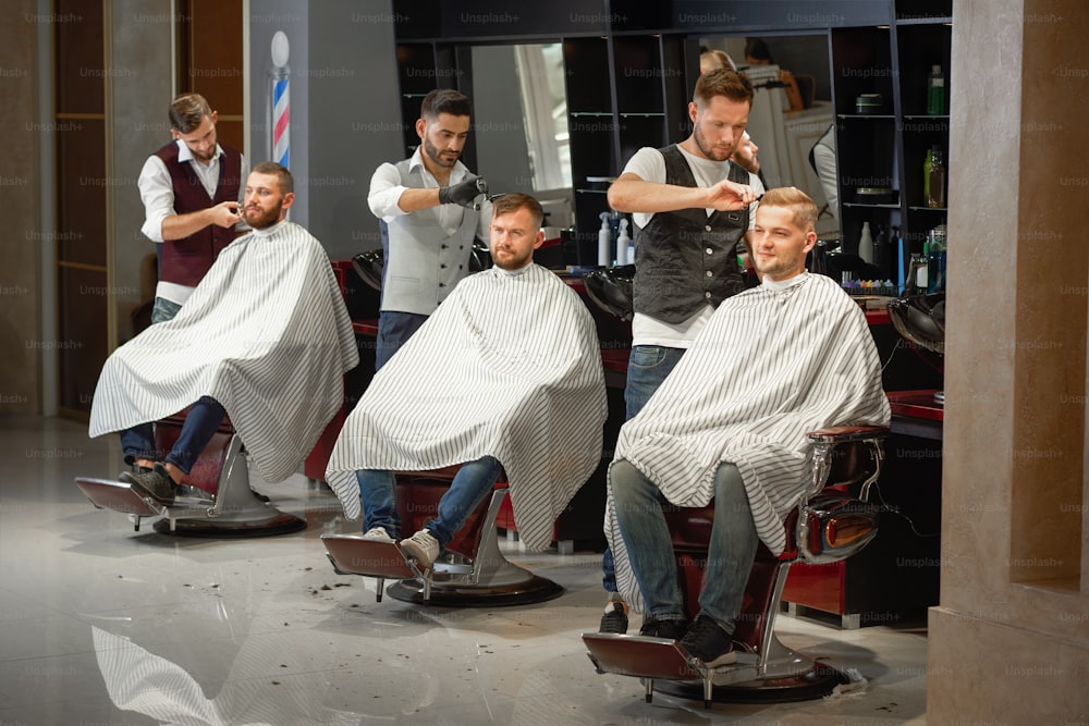 Tre barbieri in camicia bianca e gilet che acconciano capelli e barba con forbici e rasoio a mano libera nel negozio di barbiere. Giovani uomini belli seduti sulla sedia durante la procedura. Concetto di taglio di capelli.