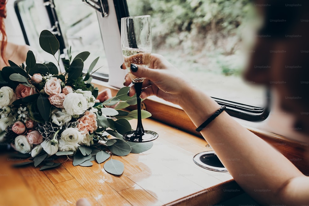 hand with glass of champagne, stylish happy bride and groom toasting and having fun with bridesmaids and groomsmen inside of retro car. emotional moment, space for text. luxury wedding