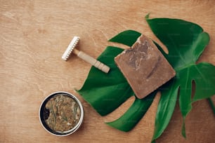Natural soap, solid eco shampoo in metal tin, reusable razor on wooden background with green monstera leaf. Plastic free beauty essentials. Zero waste concept, flat lay. Sustainable lifestyle