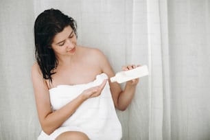Skin and body care. Young happy woman in white towel applying moisturizing cream on hand in home bathroom with green plants.  Hand holding plastic bottle with lotion. Spa and wellness