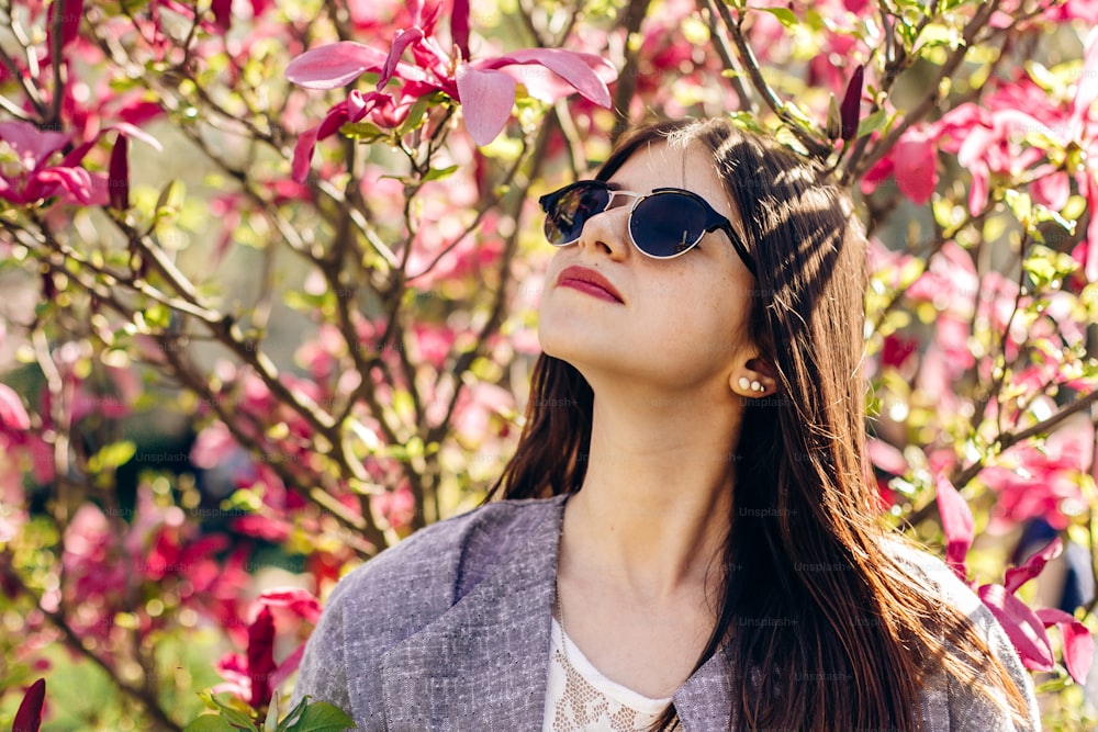 Mujer hipster con estilo disfrutando de la luz del sol en flores rosas de magnolia en un parque soleado. Joven con gafas de sol sonríe en el jardín botánico en primavera. espacio para el texto. momento de alegría.