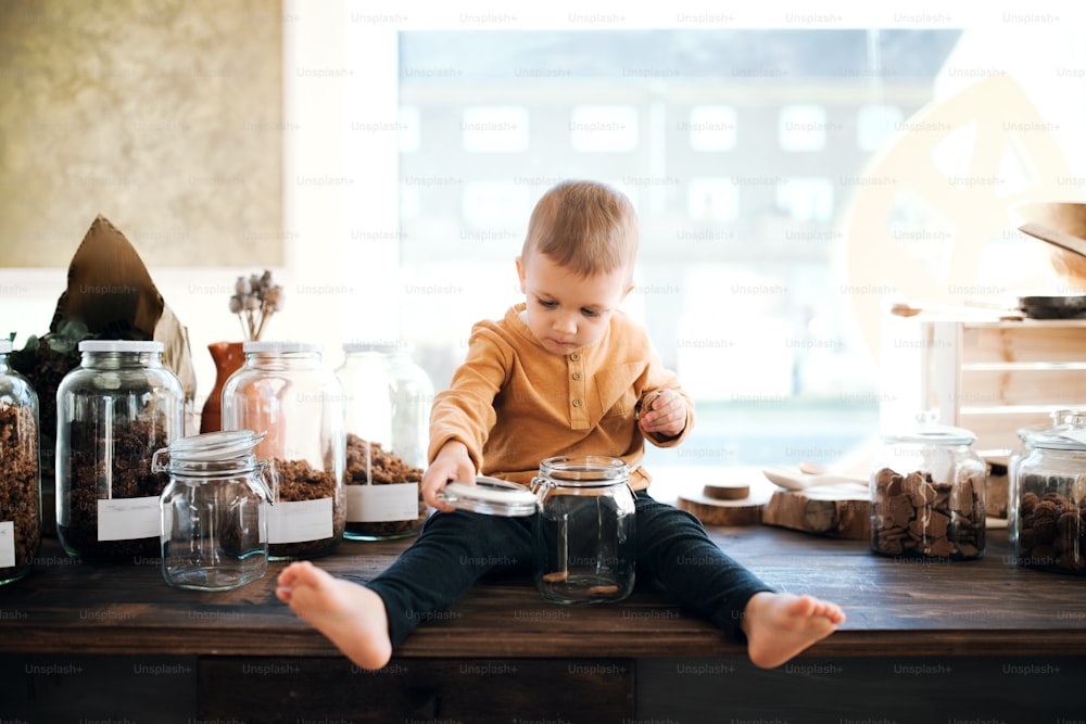 A happy barefoot small toddler boy sitting on a table in zero waste shop, holding a glass jar.