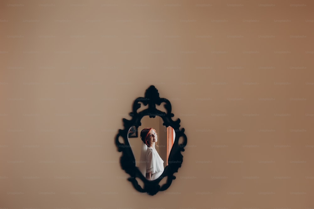 Glückliche stilvolle Braut im Seidengewand, die im Spiegel an der Wand schaut und lächelt.  Rustikale Hochzeitsmorgenvorbereitung. Porträtspiegelung einer jungen Frau. Platz für Text. Luxus-Braut