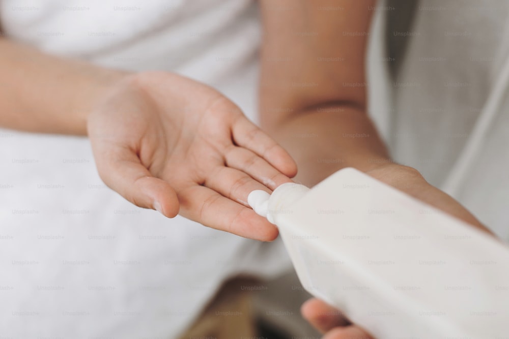 Crema hidratante de aplicación manual.  Mujer joven en toalla blanca sosteniendo botella con loción o crema en el baño. Cuidado de la piel y el cuerpo. Spa y bienestar