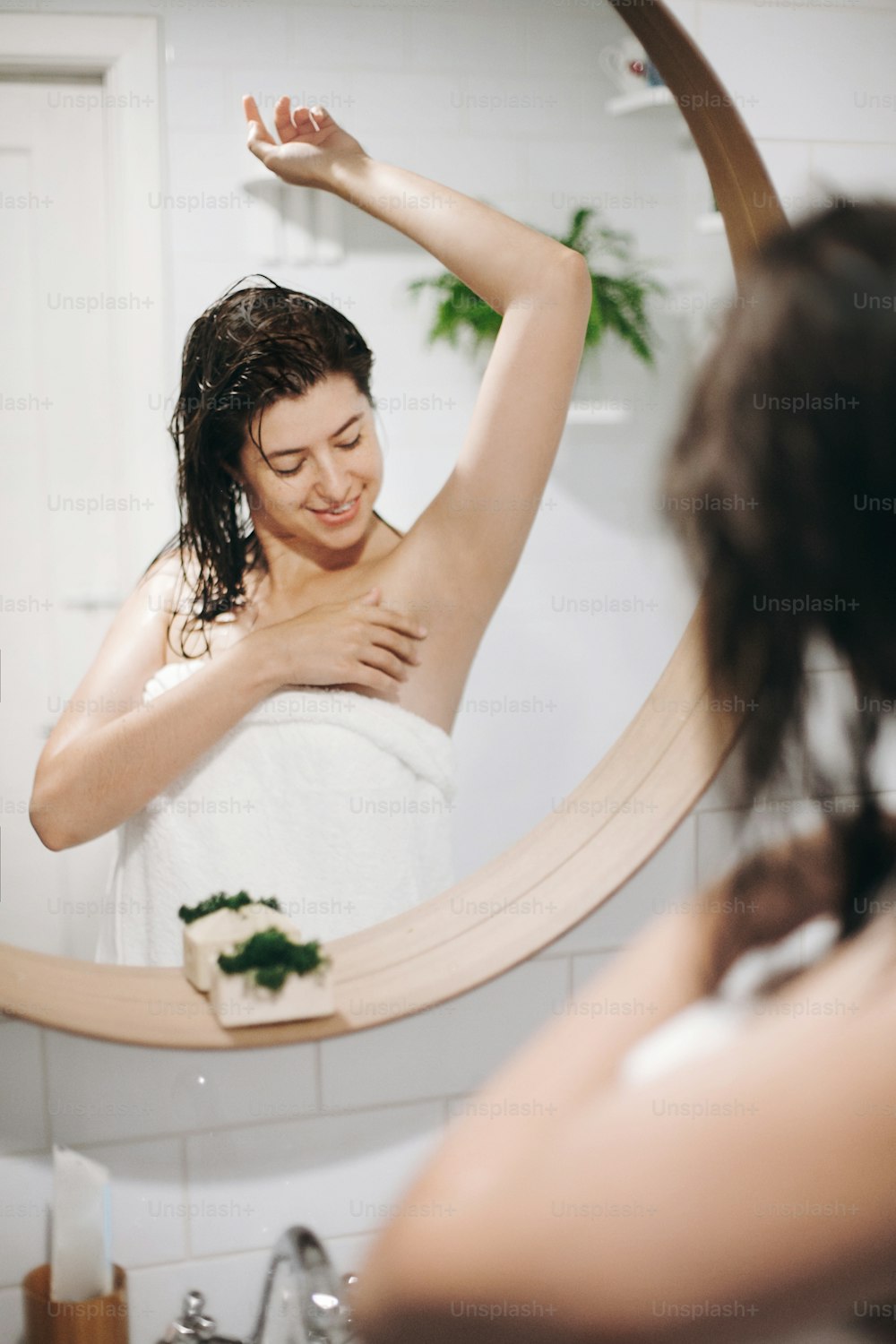 Young attractive woman with wet hair in white towel looking at  smooth soft skin after shaving armpits, reflection in mirror in stylish bathroom with greenery. Skin and body care, wellness concept