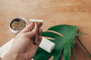 Zero waste, plastic free beauty essentials. Hand holding reusable razor for shaving on background of natural soap, solid shampoo, eco deodorant, on wood with green monstera.  Sustainable lifestyle