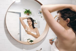 Mujer joven atractiva en toalla blanca afeitando axilas, mirándose en el espejo en un baño elegante. Cuidado de la piel y el cuerpo. Concepto de depilación. Mujer después de la ducha afeitándose con maquinilla de afeitar