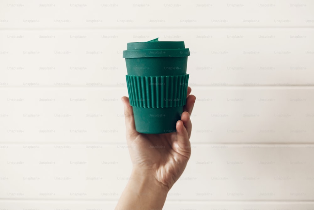 ゼロウェイストのコンセプト。白い木製の背景にスタイリッシュで再利用可能なエココーヒーカップを手に持っています。天然竹繊維のグリーンカップ。使い捨てプラスチックの禁止。選択してください。