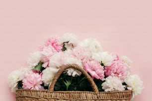 Hello spring concept. Stylish straw rustic bag with white and pink peonies on pastel pink paper, flat lay with copy space. International Women's Day. Happy mothers day, floral greeting card