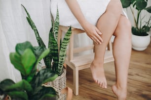 Young woman in white towel sitting in bathroom with green plants. Legs soft skin after shaving. Skin care and wellness concept.