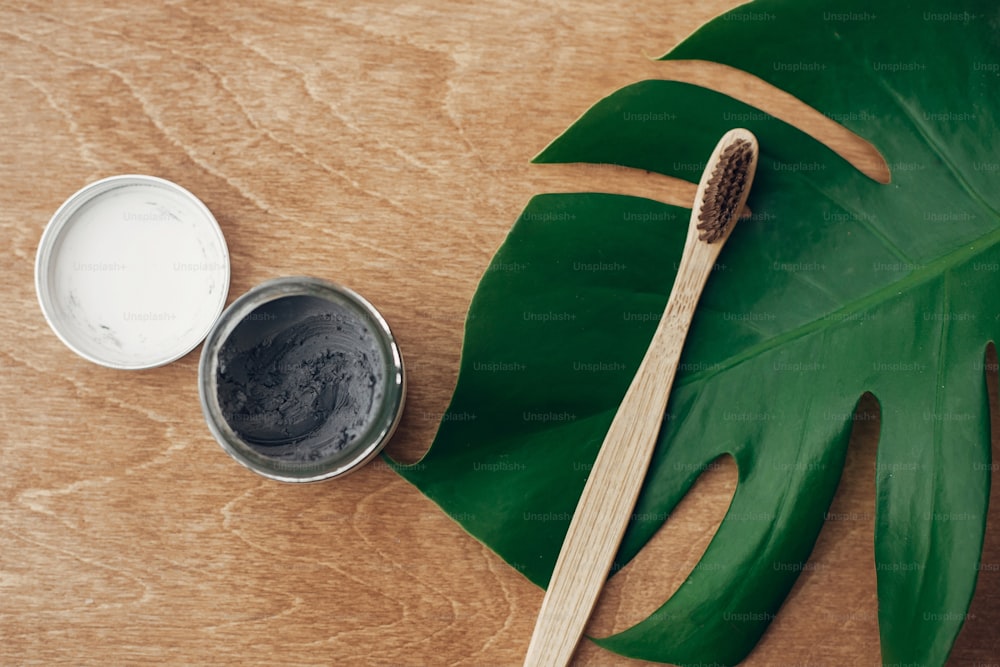 Zero waste concept. Natural toothpaste activated charcoal in glass jar and bamboo toothbrush on wooden background with green monstera leaf. Plastic free essentials, teeth care. Sustainable lifestyle
