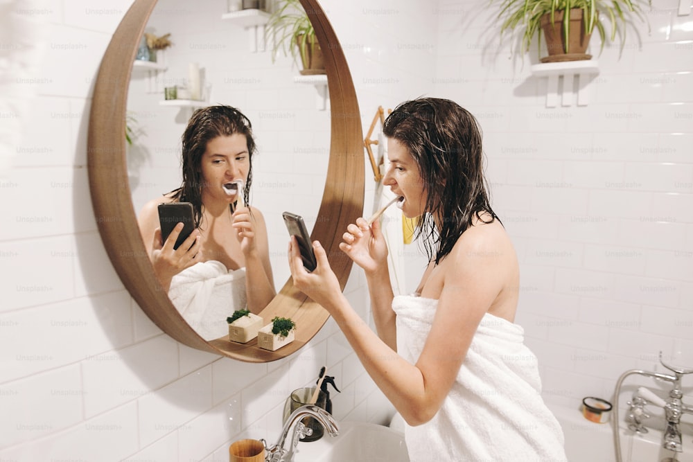 Jeune femme heureuse en serviette blanche se brossant les dents et regardant son smartphone dans la salle de bain avec miroir. Femme mince et sexy avec une peau naturelle et des cheveux mouillés, routine quotidienne après la douche. Influence des médias sociaux