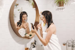 Giovane donna felice in asciugamano bianco che si lava i denti e guarda lo smartphone in bagno con specchio. Donna sexy e magra con pelle naturale e capelli bagnati routine quotidiana dopo la doccia. L'influenza dei social media