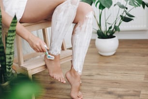 Hair Removal with depilation cream concept. Young woman in white towel applying shaving cream on her legs and holding plastic razor in home bathroom with green plants. Skin care