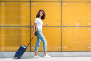Mujer usando el teléfono móvil mientras camina y empuja una maleta en un fondo de vidrio amarillo