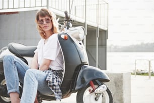 Portrait of a beautiful girl hipster sitting on a black retro scooter, smiling posing and enjoy the warm spring sunshine