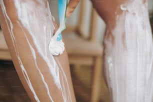 Junge Frau rasiert Beine mit Plastikrasierer Nahaufnahme und Enthaarungscreme im Badezimmer mit grünen Pflanzen. Hautpflege. Hand halten Plastikrasierer auf der Haut mit Creme. Haarentfernung