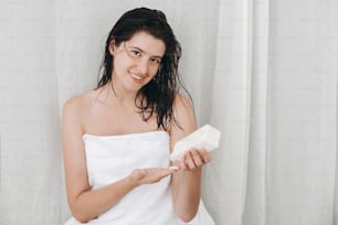 Cura della pelle e del corpo. Giovane donna felice in asciugamano bianco che applica crema idratante a portata di mano nel bagno di casa con piante verdi.  Mano che tiene bottiglia di plastica con lozione. Spa e benessere