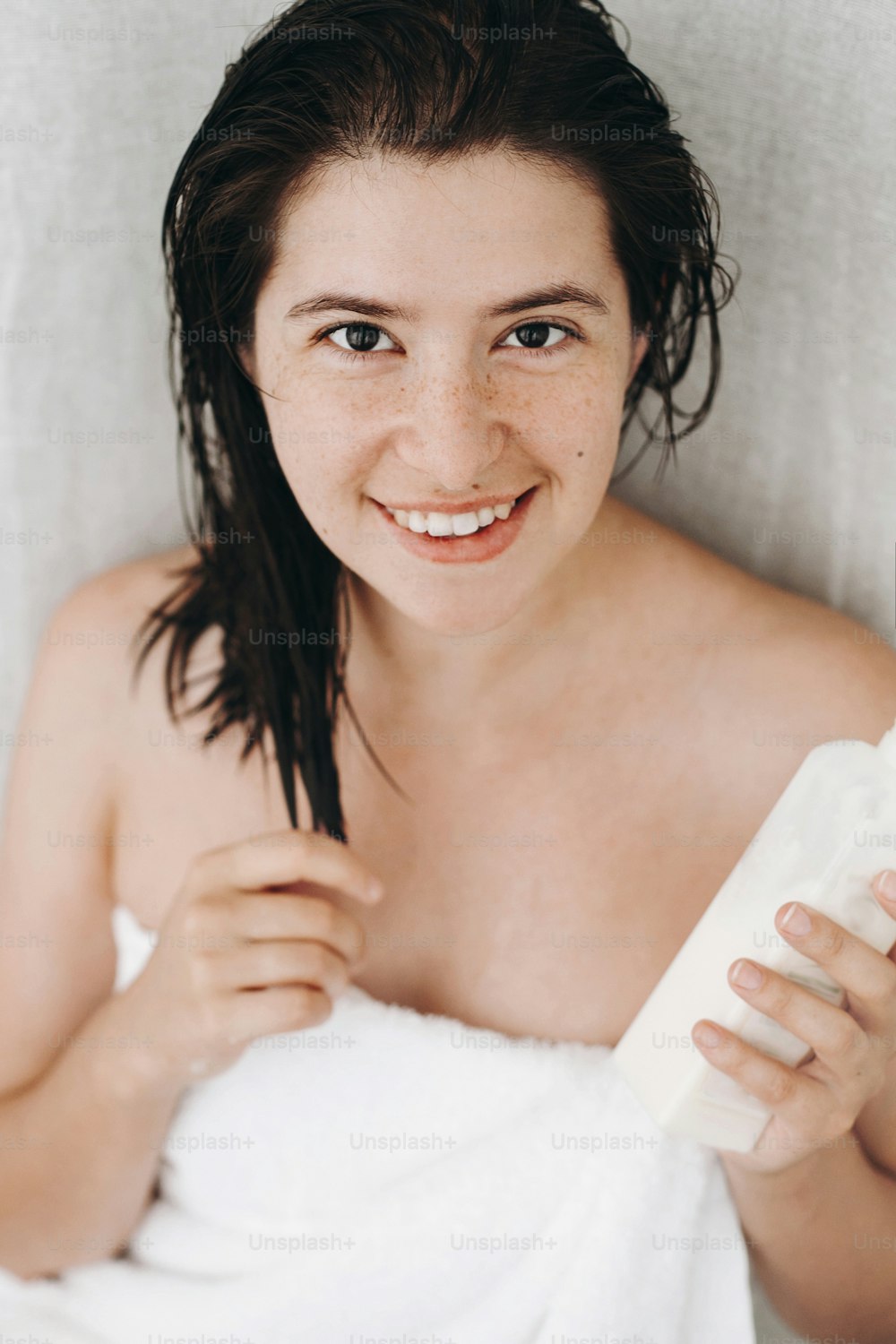 Young happy woman in white towel holding conditioner for hair and smiling in bathroom. Skin and hair care. Slim sexy woman with natural skin enjoying spa and wellness, relaxing. Copy space
