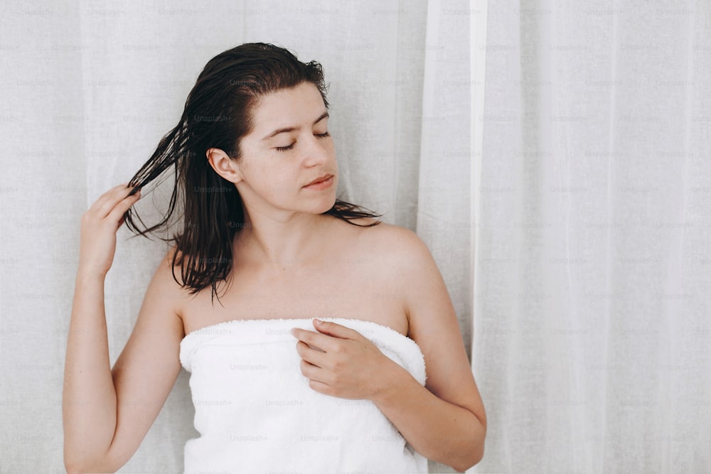 Haar- und Körperpflege. Junge glückliche Frau in weißem Handtuch, die eine Conditioner-Maske auf das Haar im Badezimmer aufträgt.  Schlanke sexy Frau mit natürlicher Haut, die Spa und Wellness genießt und sich entspannt.