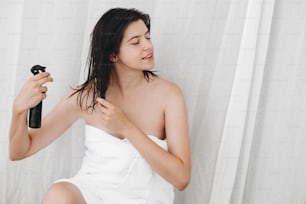 Hair and body care. Young happy woman in white towel applying conditioner mask on hair in bathroom. Slim sexy woman with natural skin enjoying spa and wellness, relaxing