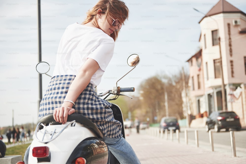 Portrait d’une belle fille hipster assise sur un scooter rétro noir, souriant posant et profitant du chaud soleil printanier