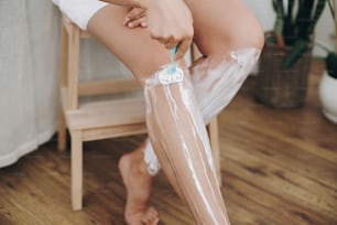 Haarentfernung mit Enthaarungscreme-Konzept. Junge Frau in weißem Handtuch trägt Rasierschaum auf ihre Beine auf und hält Plastikrasierer im Badezimmer mit grünen Pflanzen. Hautpflege