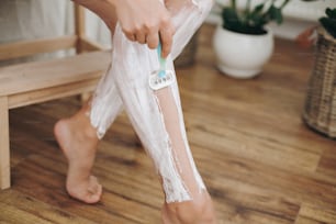 Junge Frau in weißem Handtuch rasiert Beine mit Plastikrasierer und Enthaarungscreme im Badezimmer mit grünen Pflanzen. Hautpflege. Haarentfernung mit Enthaarungscreme-Konzept.