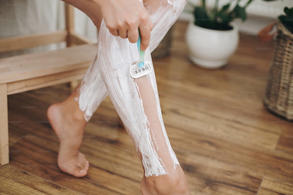 Jeune femme en serviette blanche rasant les jambes avec un rasoir en plastique et une crème d’épilation dans la salle de bain de la maison avec des plantes vertes. Soins de la peau. Épilation avec concept de crème d’épilation.