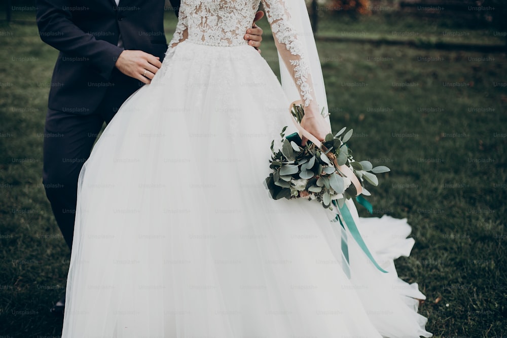 세련된 행복한 신부와 신랑이 마당을 걷고 결혼식에서 손을 잡고 있다. 낭만적 인 순간, 텍스트를위한 공간. 럭셔리 웨딩 신혼 커플, 꽃다발과 가운 클로즈업