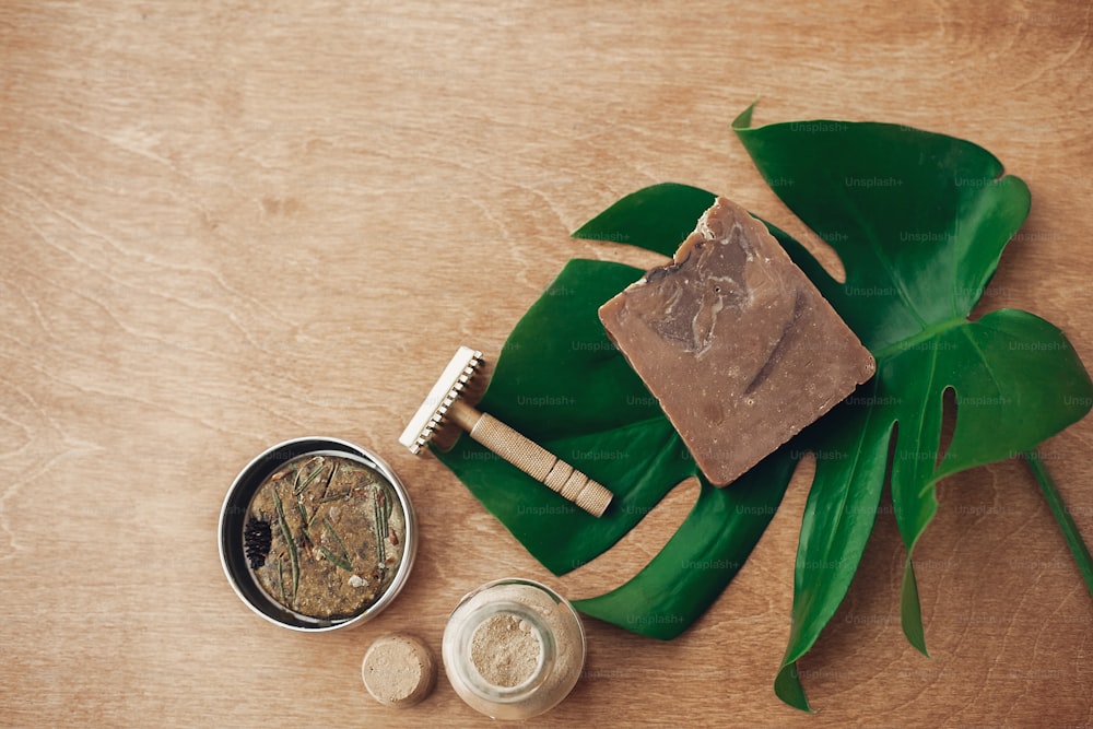 Natural soap, solid shampoo in metal tin, reusable razor, ubtan on wooden background with green monstera leaf. Plastic free beauty essentials. Zero waste concept, flat lay. Sustainable lifestyle