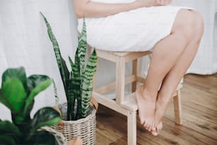 Young woman in white towel sitting in bathroom with green plants. Legs soft skin after shaving. Skin care and wellness concept.
