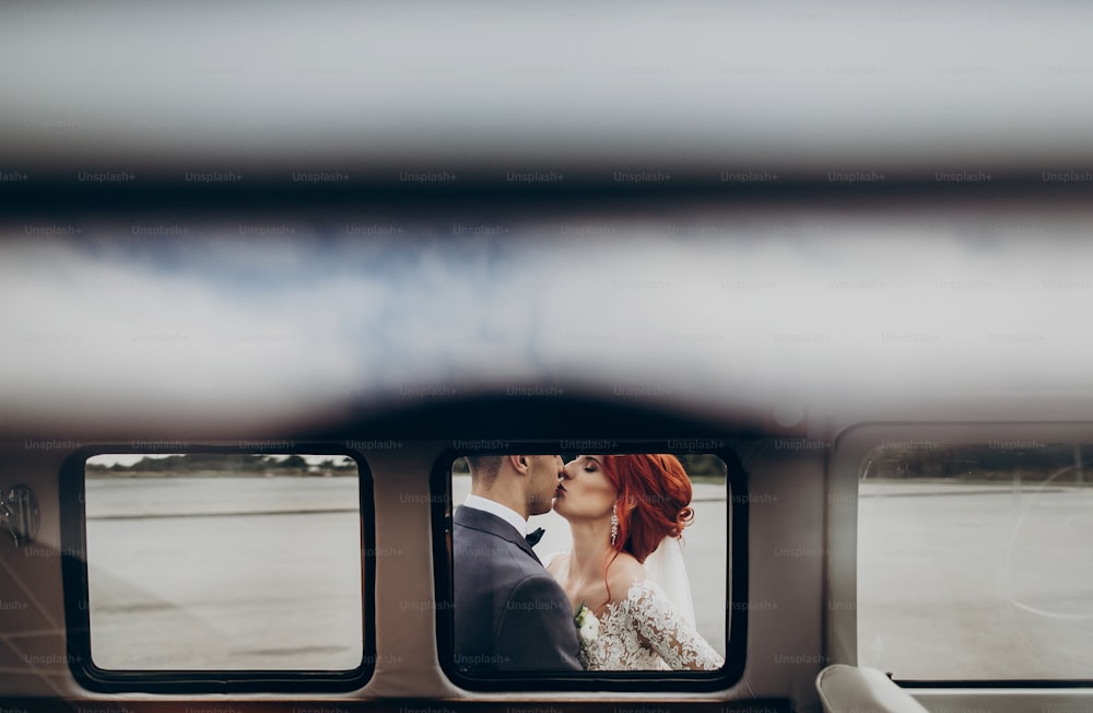 Stilvoller Bräutigam und glückliche Braut umarmen und küssen sich sanft im Retro-Auto. Emotionaler romantischer Moment, Raum für Text. Ungewöhnlicher Blick durch Fensterauto bei Luxushochzeit Brautpaare
