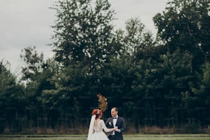 stylish bride and groom walking and smiling on background green trees, holding hands. luxury wedding couple newlyweds dancing, happy emotional moment. space for text. true feelings