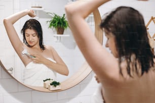 Mujer joven atractiva en toalla blanca afeitando axilas, mirándose en el espejo en un baño elegante. Cuidado de la piel y el cuerpo. Concepto de depilación. Mujer después de la ducha afeitándose con maquinilla de afeitar