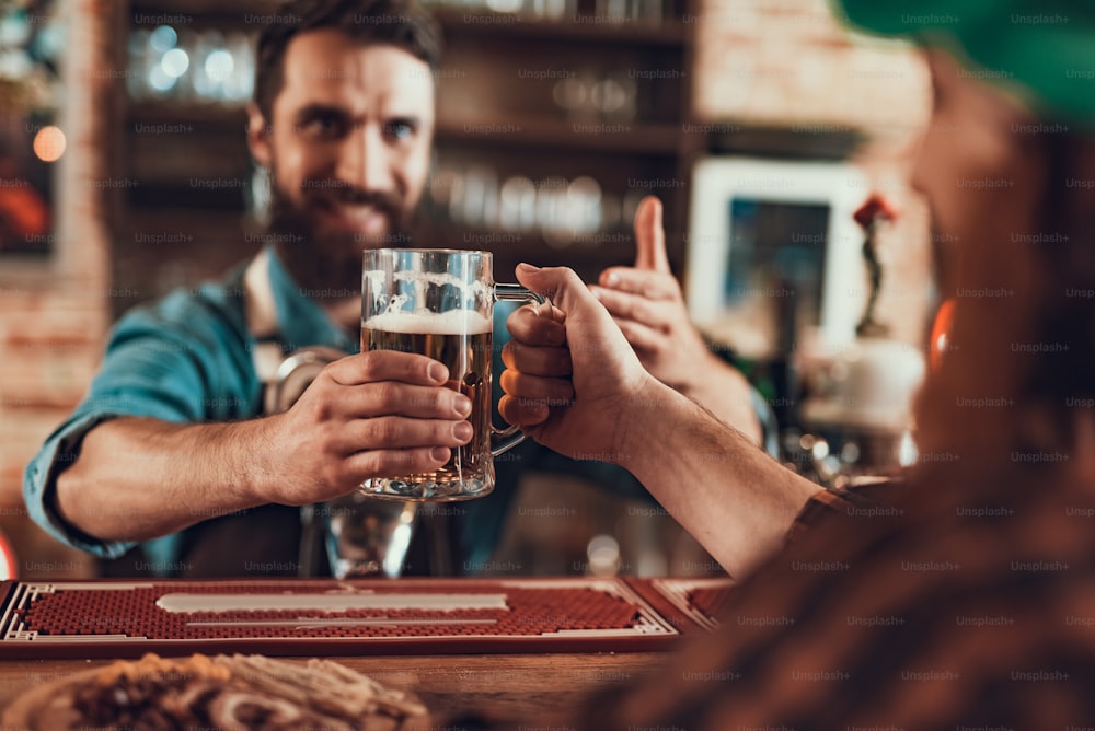 Primer plano de manos masculinas sosteniendo una jarra de cerveza fresca con espuma. Barman barbudo sonriente sobre fondo borroso