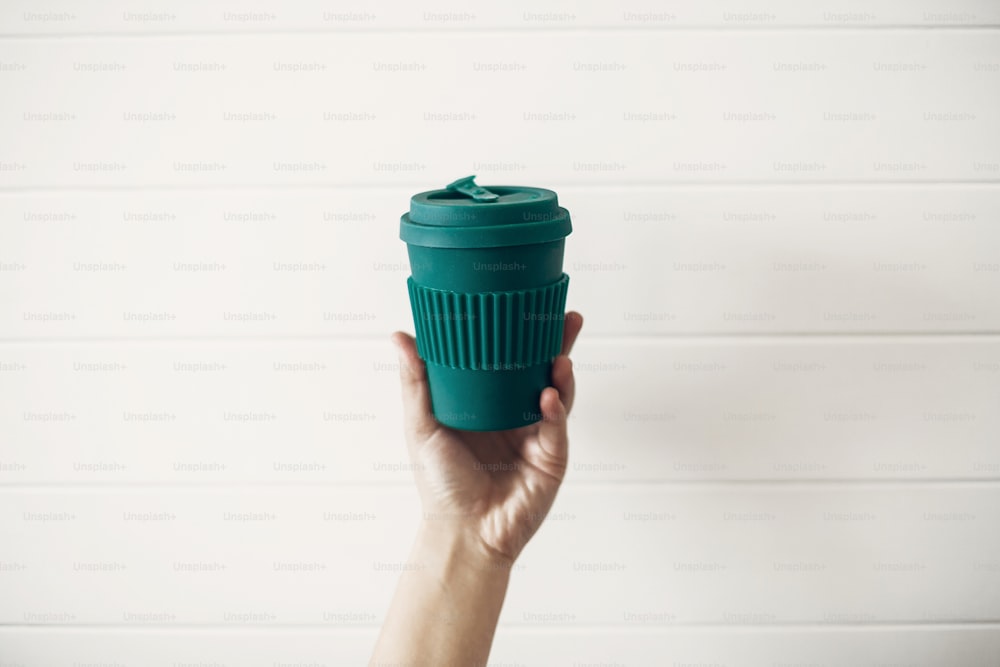 Hand hält stilvolle wiederverwendbare Öko-Kaffeetasse auf weißem Holzhintergrund. Green Cup aus natürlicher Bambusfaser, Zero-Waste-Konzept. Verbot von Einwegplastik. Treffen Sie eine Wahl. Kaffee zum Mitnehmen